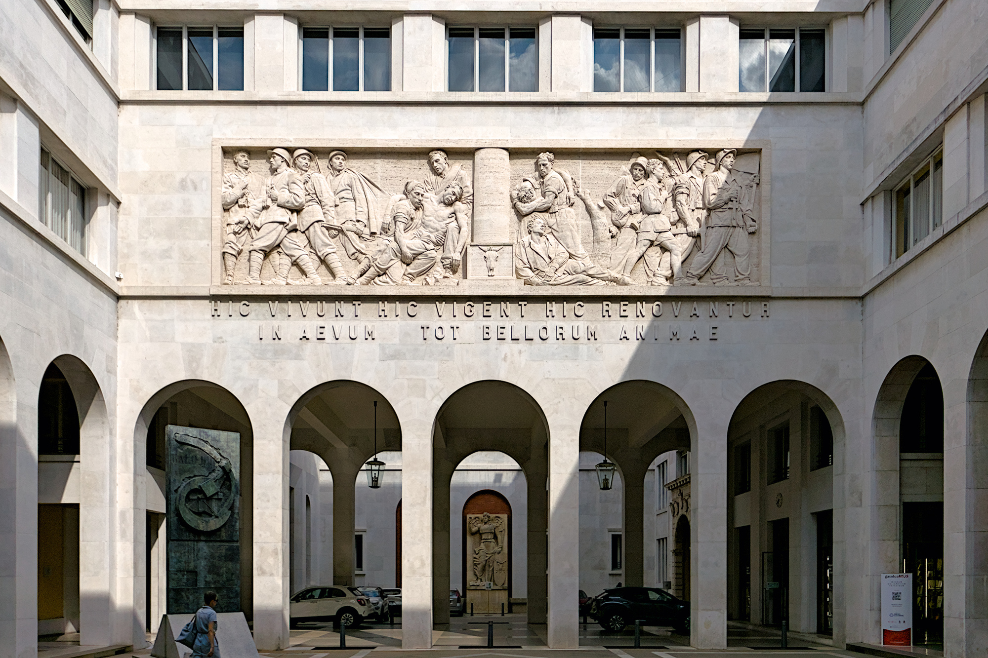 Attilio Selva, *Elogio al coraggio degli studenti in battaglia*, altorilievo, 1939. In lontananza, sulla parete in fondo: Paolo Boldrin, l'ateneo di Padova rappresentato come *Minerva vittoriosa*, 1942. Sulla sinistra: Giò Pomodoro, *Spirale per Galileo Galilei*, stele in bronzo (originariamente collocata in via VIII Febbraio, davanti al municipio), 1992. Via San Francesco, Padova - palazzo Bo, Cortile nuovo (in origine 'Cortile littorio')